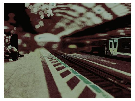 Das Bild zeigt eine Szene in einer U-Bahn-Station. Im Vordergrund ist eine Gleislinie zu sehen, die in die Ferne führt, und ein Teil des Bahnsteigs mit einem markierten Sicherheitsstreifen in Weiß und Grün. Auf der linken Seite sitzen zwei Personen auf dem Bahnsteig, eine davon hat einen Rucksack. Rechts fährt ein Zug an, dessen Türen offen sind. Die Hintergrundbeleuchtung erzeugt eine bokeh-Effekt mit verschwommenen Lichtern, die eine dynamische Atmosphäre schaffen. Die Farbgebung des Bildes ist sepia-ähnlich, was eine nostalgische Stimmung vermittelt.