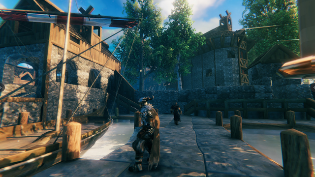Valheim Screenshot: In der Mitte ein Mann in Rüstung, Fellkleidung, Helm und Umhang steht an einem Kai. Links ist ein Segelschiff mit aufgerollten rot-weiß gestreiften Segeln zu sehen. Weiter hinten mehrere steinerne Gebäude und Laubbäume.