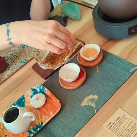 Close up of a hand pouring tea from a glass pitcher to a small ceramic cup. A small white teapot is visible nearby, and a card indicates that the tea is Dong Ding taiwanese oolong.