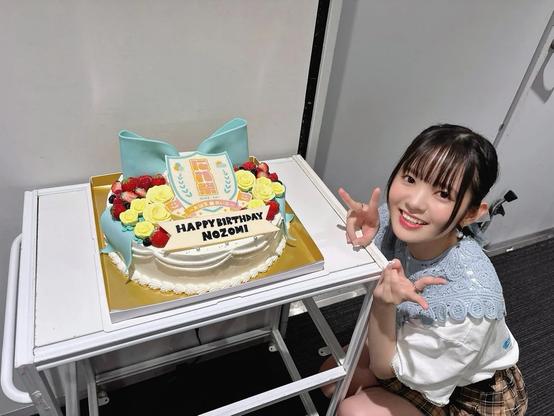 A young woman is smiling and posing with a peace sign next to a beautifully decorated birthday cake. The cake is adorned with yellow roses, strawberries, a blue ribbon, and a sign that reads “HAPPY BIRTHDAY NOZOMI.” She appears happy and cheerful, celebrating a special occasion in a casual indoor setting.