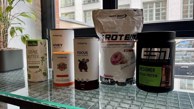 The image features various protein supplements arranged on a table. From left to right, there is a green can labeled "Grüne Mutter" by Alpha Foods, a white container of whey protein from foodspring, an orange tub of "Energy Aminos"