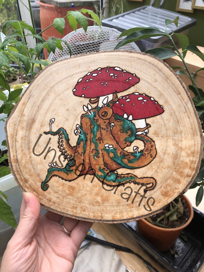 Roundish wood slice with burned and painted design showing an orange octopus sitting underneath red and white amanita mushrooms. He has tiny white mushrooms growing on him and green places that could be moss or disease, who knows 🤷‍♀️