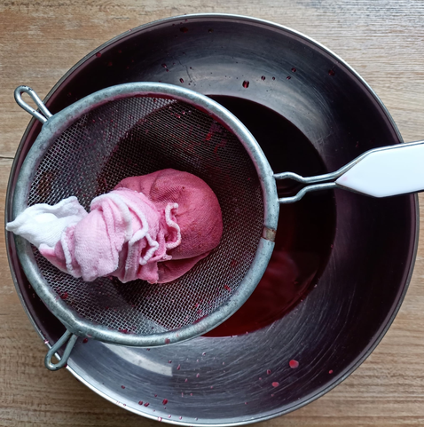Eine Schüssel, darin der fertige Himbeer-Essig. Darüber ein Sieb mit einem zusammengedrückten Tuch, das vom Himbeer-Essig rosa eingefärbt ist.