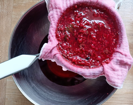 Eine Schüssel, darüber ein Sieb mit einem feinen Tuch, darin die rosa- Masse aus Himbeeren, Essig und Gewürzen