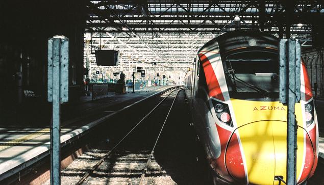 A view inside Edinburgh Waverley looking down the platform from the buffer stops. On the right is a red and white Azuma train, with a yellow front. In the foreground the platform on the left is dark but in the background there is a lot of light coming from the glass roof
