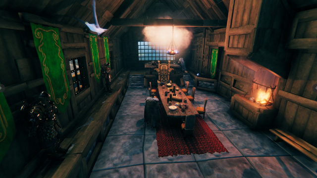 Valheim Screenshot: Ein Saal mit Steinfußboden und Holzwänden. In der Mitte ein langer Tisch, auf dem Geschirr, Brot und Goldmünzen liegen. Am Kopfende des Tisches steht ein hölzerner Thron, die restlichen Sitzgelegenheiten sind schlichter. An den Wänden hängen grüne Banner und es sind zwei Rüstungen aufgestellt. Auf der rechten Seite brennt ein Kaminfeuer.
