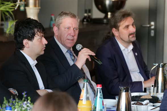 Das Bild zeigt eine Gesprächsszene beim 2. Parlamentarischen Frühstück der Cyberagentur. Im Vordergrund sitzen vier Männer nebeneinander an einem gedeckten Tisch mit Kaffee- und Wasserflaschen, Tassen, Gläsern und einem Brotkorb. In der Mitte des Bildes spricht ein Mann mit grauen Haaren und Anzug in ein Mikrofon – es handelt sich um Andreas Schwarz, MdB, der hier offenbar einen Redebeitrag leistet. Links neben ihm sitzen zwei Männer mit dunklen Haaren in dunklen Anzügen, sie hören aufmerksam zu. Rechts neben ihm sitzt ein weiterer Mann mit längeren Haaren und Bart, ebenfalls im Anzug, der mit leichtem Lächeln dem Geschehen folgt. Im Hintergrund sind Pflanzen, Getränkeflaschen und metallene Gefäße zu sehen. Die Stimmung wirkt konzentriert, aber offen, wie bei einer konstruktiven Diskussion am Frühstückstisch in einem formellen Rahmen. Foto: Cyberagentur
