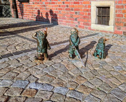 Three bronze statues of Wrocław Dwarfs stand on a cobblestone street. The dwarf on the left, representing deafness, wears a pointed hat and puts a hand to his ear, looking upwards. The dwarf in the middle, representing blindness, holds a white cane. The dwarf on the right is in a wheelchair. A red brick building with a window is visible in the background. The shadows of people are cast on the building.