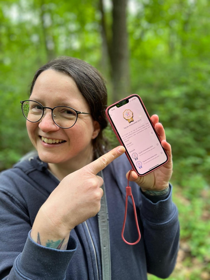 Eine lächelnde Frau mit blauem Kapuzenpulli und Brille zeigt auf ein Telefon, welches das Abzeichen präsentiert.