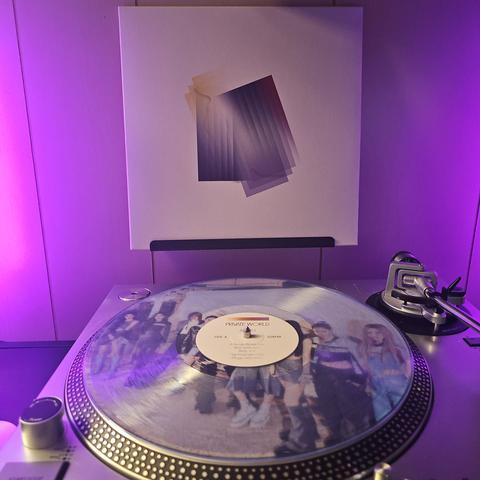 A clear vinyl record sits on a turntable. Behind the turntable, a vinyl album outer sleeve is displayed. The front cover shows multiple gradient rectangles overlapping each other.