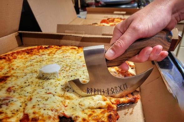 Photo of a hand holding a mini-axe pizza cutter slicing a thin-crust (tavern style) square-cut pizza. A white pizza stand is visible in the middle and another pizza is in the background.  The axe has letters written on the side near the blade