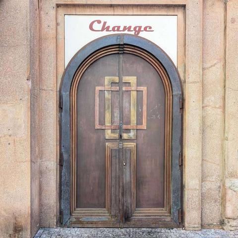 An arched double door with geometric brass inlays, set in a weathered stone facade. Above the door, a white rectangular sign with the word "Change" written in red cursive is visible. The doorway is framed by dark metal trim, and the ground in front is paved with cobblestones.