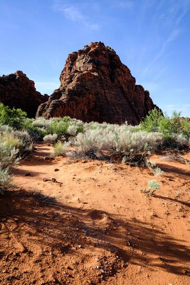A rocky desert landscape with a large red rock formation in the background, sparse green shrubs, and sandy ground in the foreground under a clear sky.