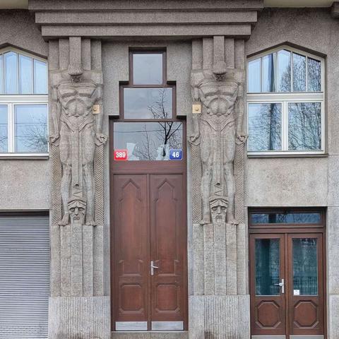 A street-level view of an Art Deco building facade featuring a prominent wooden double door entrance. Flanking the doorway are two massive, carved stone figures of muscular men supporting the architectural structure above. Directly above the main door are the numbers "389" in a red square and "46" in a blue square. To the right of the main entrance is a smaller wooden door. Large arched windows are visible on either side of the central doorway. The building is constructed of gray stone with vertical striations.