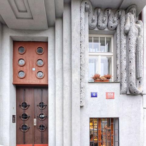 A street-level view of an Art Deco building facade with a striking brown double door on the left. The door features circular glass insets. The building is constructed of light gray concrete with strong vertical lines and geometric shapes. To the right of the door, a window is framed by stylized, elongated stone figures with circular patterns. Blue and red plaques display the numbers "6" and "1556" respectively. A smaller window is visible below the larger one, with decorative metalwork.
