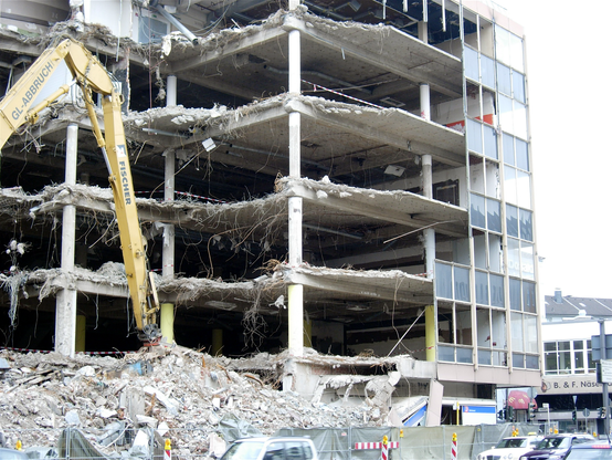 geschäftshaus während des abbruchs … die fassade ist schon weg … man kann ins innere sehen … ein gewirr aus kabeln hängt herum … im vordergrund der arm eines baggers und ein schuttberg