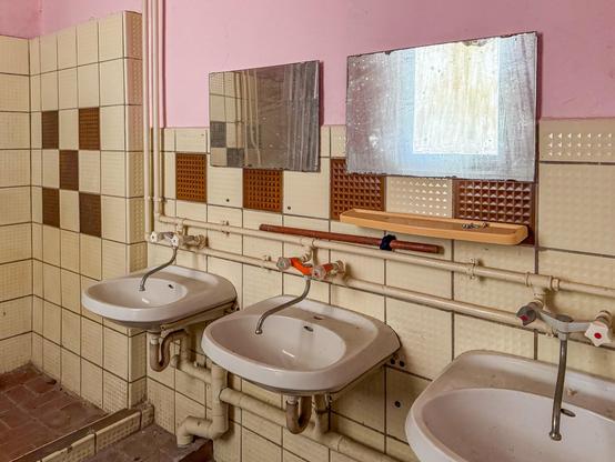 Verlassener, ehemaliger Waschraum, Vordergrund sind zwei Spiegel vor einer gefliesten Wand zu sehen, die Fliesen sind beige, es gibt einige wenige, akzentuierte braune Fliesen, alle Wasserleitungen sind beige, gestrichen und über Wand und Fliese installiert, im Vordergrund sind drei Waschbecken mit den dazugehörigen Wasserhähnen und den Abflussleitung zu sehen, da drüber sind die zwei Spiegel, durch die man schemenhaft auf der anderen Seite des Raumes ein schmutziges Fenster erkennen kann