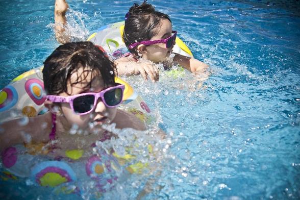 Two children are playing in a swimming pool, wearing bright sunglasses and enjoying the water. One child is floating on a colorful inflatable, while the other is splashing nearby. Sunlight reflects off the water, creating a lively, fun atmosphere.