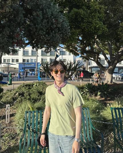 Smiling SUHO wearing a pale green tee and a rose colored kerchief tied around his neck to keep the sweat off. He stands in front of a pair of green metal benches, bushes and a tree behind him. Across the street behind the tree is a Ghirardelli storefront and a Fogo de Chao storefront.