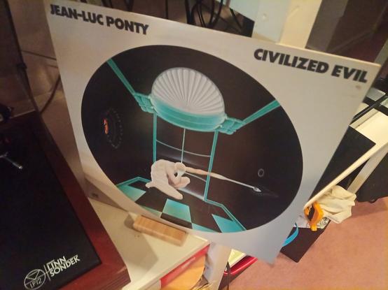 The image shows an album cover for "Civilized Evil" by Jean-Luc Ponty. The cover features a circular design with a dark background. In the center, there is a white figure resembling a rabbit, sitting on a checkered floor with teal lines. Above the figure, there is a teal dome-like structure with a fan-like pattern. The text "JEAN-LUC PONTY" is at the top, and "CIVILIZED EVIL" is at the bottom. The album is placed on a table with a Linn Sondek turntable visible on the left side. The overall color scheme includes black, white, teal, and hints of red.

Provided by @altbot, generated privately and locally using Ovis2-8B