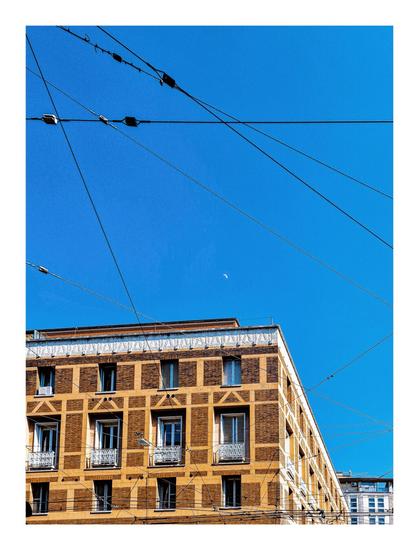 This is a photo of an upper section of a multi-story brick building against a bright blue sky. There are lots of overhead wires, used for buses, that crisscross the frame. The intersect with the building's facade. A sliver of a crescent moon is visible in the upper portion of the sky near the middle of the photo frames by the wires. There is the sliver of a second building behind the first. It is grey with lots of windows.