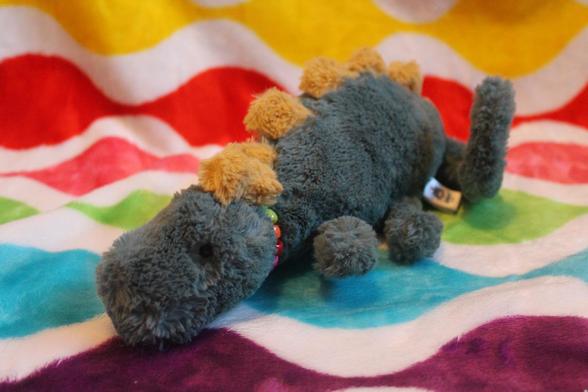 A little dark green dinosaur plush with yellow spikes along his spine, placed on a backdrop made with a colourful rainbow blanket. He's positioned in a lying down position, with a long tail curled around he's body. He's also wearing a rainbow kandi necklace.