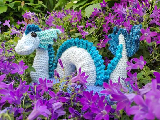 Dragon de mer au crochet immergé dans un socle bleu représentant la mer, au milieu des violettes. Le dragon est bleu clair, avec des nageoires et une crête en dégradé de bleu.