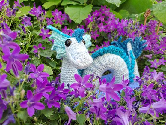 Dragon de mer au crochet immergé dans un socle bleu représentant la mer, au milieu des violettes. Le dragon est bleu clair, avec des nageoires et une crête en dégradé de bleu. Vue légèrement de côté.