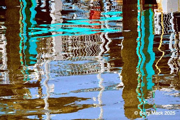 Colorful water abstract photograph of mostly vertical forms reflected in slightly moving water. The forms are of various thicknesses and colors incluing brown, turquoise, yellow, light blue, red, and green. Where it can be seen, the water is blue.