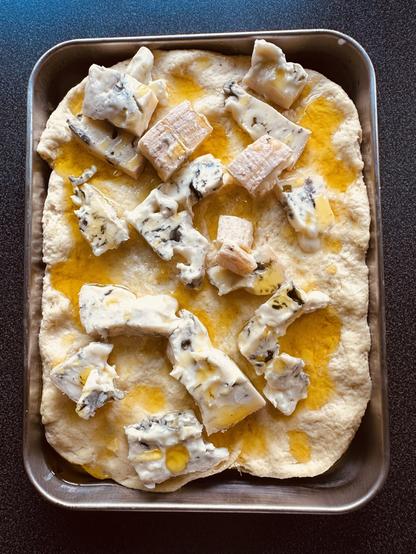 A rectangular focaccia topped with chunks of blue cheese and drizzled with olive oil is set on a baking tray.