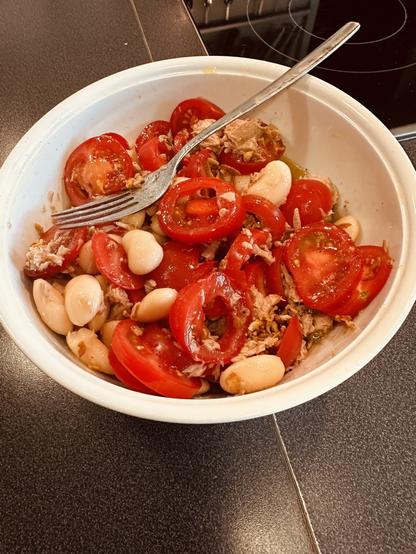 A bowl contains a salad made of sliced tomatoes, white beans, tuna, and dressing, with a fork placed on the side.