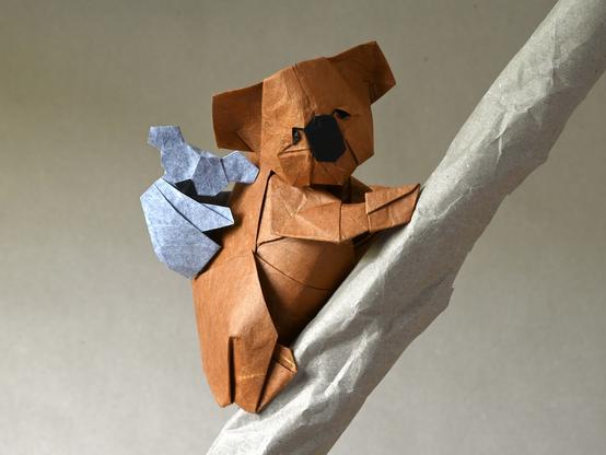 A realistic looking origami Koala with a smaller, simpler one on its back. The big one is sitting on a twig also made of paper.