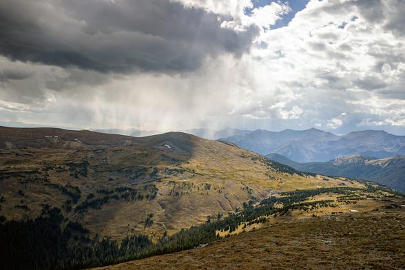Rain falling from dark clouds over mountains. But is it reaching the ground? Scenic photo in Rocky Mountain National Park, looking west from Trail Ridge Road over Forest Canyon toward the Never Summer Range. Dark gray clouds above a shadowed mountain ridge at left, with streaks of rain falling that may or may not be reaching the ground. The right half of the image is sunlit valley with a range of mountains in the background, bright white clouds above. The air is typically very dry and rain often falls but doesn’t reach the ground. It’s called virga. Not to be confused with verga, which means penis in Spanish. September 2004.
