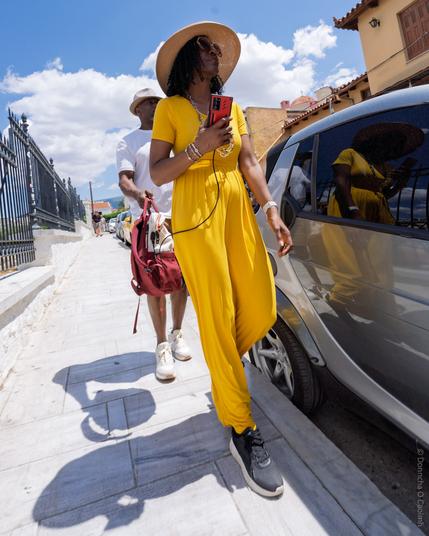 A candid street photo taken in Athens, Greece, showing a stylish woman in a vibrant yellow jumpsuit and large brim straw hat walking on a sunny day, holding a red phone and headphones, with a man carrying a red bag behind her; her reflection is visible in a parked car beside them, and the scene is set against a backdrop of a wrought iron fence, white pavement, and Mediterranean architecture under a blue sky with scattered clouds.