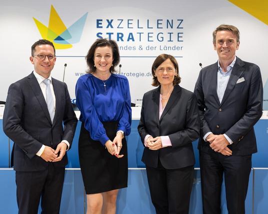 Vier Personen stehen vor einer Wand mit dem Schriftzug 'Exzellenzstrategie des Bundes und der Länder', wobei zwei Männer Anzüge tragen und zwei Frauen in Business-Outfits gekleidet sind.