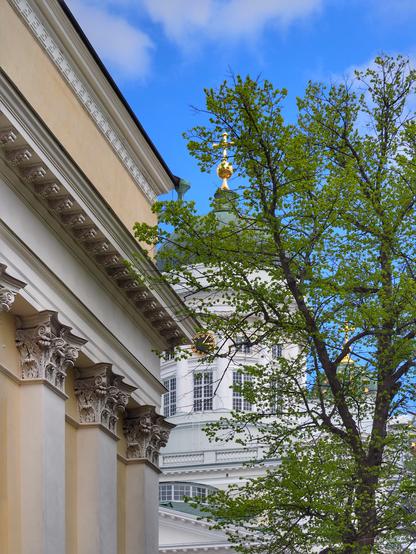 A close-up view of architectural details from a historic building, featuring ornate columns with floral designs, and a tree with fresh green leaves. In the background, a dome with a golden spire is visible against a blue sky with scattered clouds.