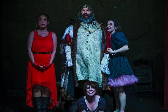 Color photo. Four people in vaguely medieval costumes. The middle is a large bearded man wearing a crown. The others are women: on the left wearing a red dress, on the right, a green dress with pink fringe at the bottom. The man is standing over the third woman, who is lying on the stage, facing the viewer, propped up on her elbows, smiling.