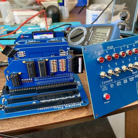 Photograph of the assembly process. The main backplane is pictured with a few modules connected, and the front panel is propped up nearby being tested. Multimeter and soldering iron in the background.