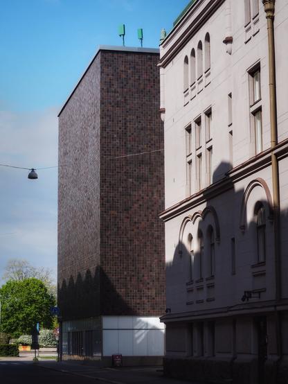 An urban scene featuring a modern dark brick building on the left and a classical light-colored building on the right. The sky is clear with some clouds, and greenery can be seen in the background. Shadows create interesting patterns on the buildings.