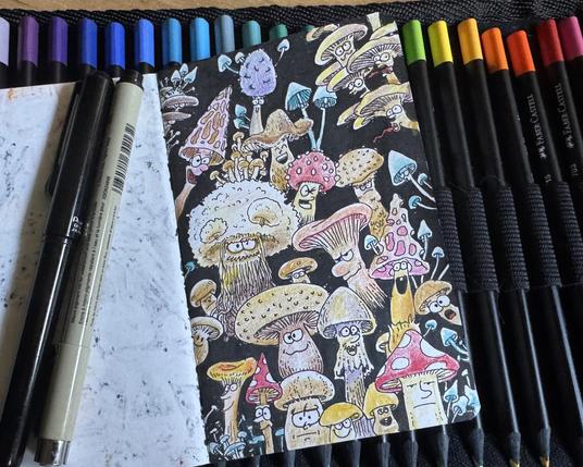 Photo of a sketchbook page with a drawing of cartoonish mushrooms in different shapes and facial expressions. Illustration is on black ink and coloured with colour pencils. A marker and a brush pen sit on the left page and a row of colour pencils under the sketchbook.