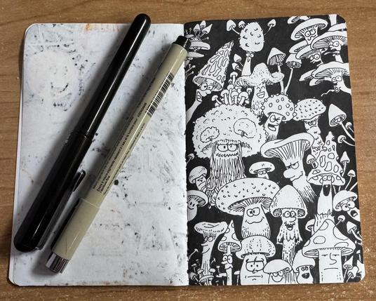 Photo of a sketchbook page with a drawing of cartoonish mushrooms in different shapes and facial expressions. Illustration is made just on black ink. A marker and a brush pen sit on the left page.
