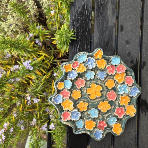 Et lavt fad med bølget kant, fyldt med keramikblomster i orange, rød og blå i forskellige højder, fyldt med vand så der hele tiden er tørre steder bier kan sidde og drikke. Det står på et sort bord, lige ved siden af en kasse med rosmarin der blomstrer