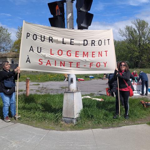 Au coin de deux rues, deux personnes tiennent une grande banderole où est écrit "Pour le droit au logement à Sainte-Foy" . En arrière, un terrain vague où d'autres personnes s'affairent, avec ce qui semble être d'autres affiches.
