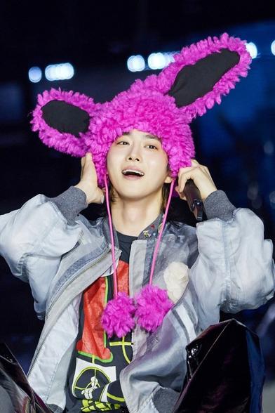 SUHO has a big happy smile, sitting on the stage, as he adjusts a bright pink fluffy hat on his head. The hat has two huge skewed rabbit ears and two dangling strings with bright pink pom poms at the ends.