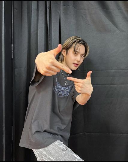 SUHO backstage in front of a gray curtain. He wears a similar gray t-shirt that had the "Welcome to SU:HOME" logo script on it. A ring dangles on a necklace around his neck. He makes two L shapes with both hands, gesturing towards the camera.