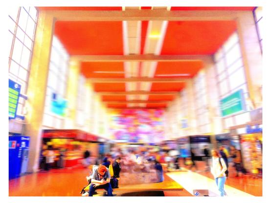 Das Bild zeigt eine verschwommene Darstellung eines großen, hellen Innenraums, der wahrscheinlich ein Bahnhof oder ein Flughafen ist. Der Raum hat eine helle, offene Architektur mit hohen Fenstern, die viel natürliches Licht hereinlassen. Die Decke ist in einem leuchtenden Orange gehalten, mit weißen Streifen, die die Deckenleuchten betonen. Im Hintergrund sind farbenfrohe Wandkunstwerke zu sehen, die dem Raum eine lebendige Atmosphäre verleihen.

Im Vordergrund sitzt ein Mann mit roten Haaren auf einer Bank und scheint auf seinem Smartphone zu arbeiten. Er trägt eine graue Jacke und eine Sonnenbrille. Neben ihm sitzt eine Frau mit dunklem Haar, die ebenfalls auf ihrem Smartphone konzentriert ist. Im Hintergrund sind weitere Menschen zu sehen, die sich bewegen oder stehen, was auf eine lebendige Umgebung hinweist.

Auf der linken Seite des Bildes sind Informationsschilder und Automaten zu erkennen, die typisch für solche öffentlichen Räume sind. Die rechte Seite zeigt weitere Geschäfte oder Informationsschalter. Die gesamte Szene ist durch die Bewegung der Menschen und die verschwommene Kameraführung dynamisch und lebendig.