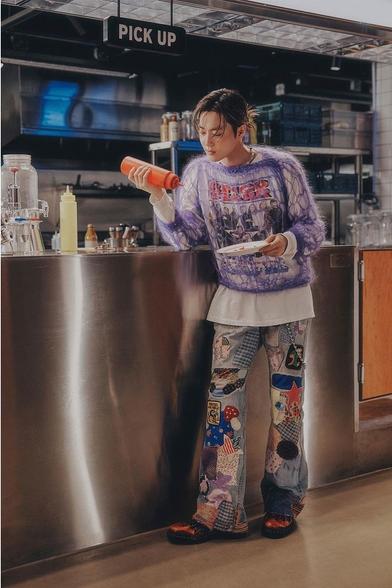 SUHO stands at a stainless steel diner counter, holding a red squeeze bottle of ketchup. He wears colorfully patched bell bottom pants, a long sleeved white Ringo Starr t-shirt, and a purple open-knit sweater on top. His hair is back in a ponytail.
