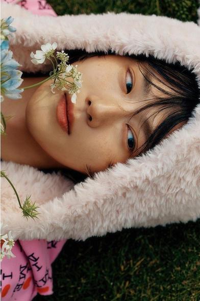 A soft, close-up portrait of SUHO's face. He lies on green grass, wearing a fluffy pale pink hood (with bunny ears that are off camera), holding some delicate flowers near his face. His eyes are looking far to his left.