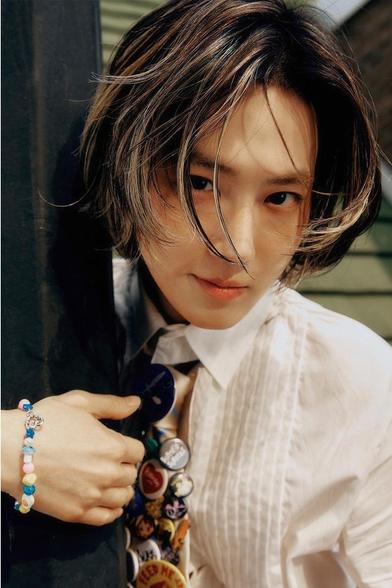 SUHO hugs a black metal sign post. The arm hugging it has a pastel beaded fan bracelet. His shirt is a white pleated tuxedo shirt. His necktie is completely covered with various sized and colored slogan buttons. He looks at the camera up through his cheek-length bangs.