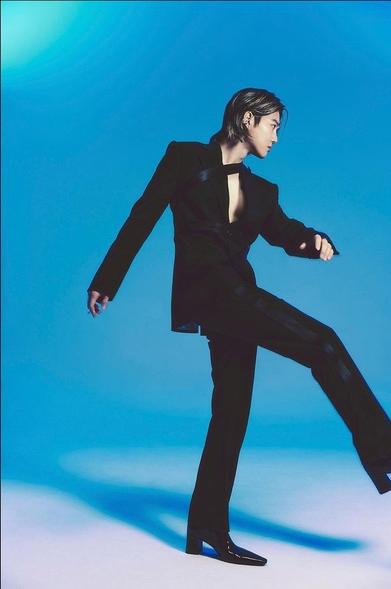 SUHO struts in a long, solid black suit, with shiny black stripes along the outside of the pant legs. His black shiny boots have tall, skinny, square heels and long narrow pointed toes. He is looking away and doing a strut-like kick with one leg.